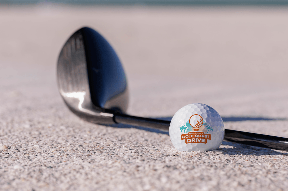 golf ball and club on the beach sand with the golf coast drive logo on the golf ball