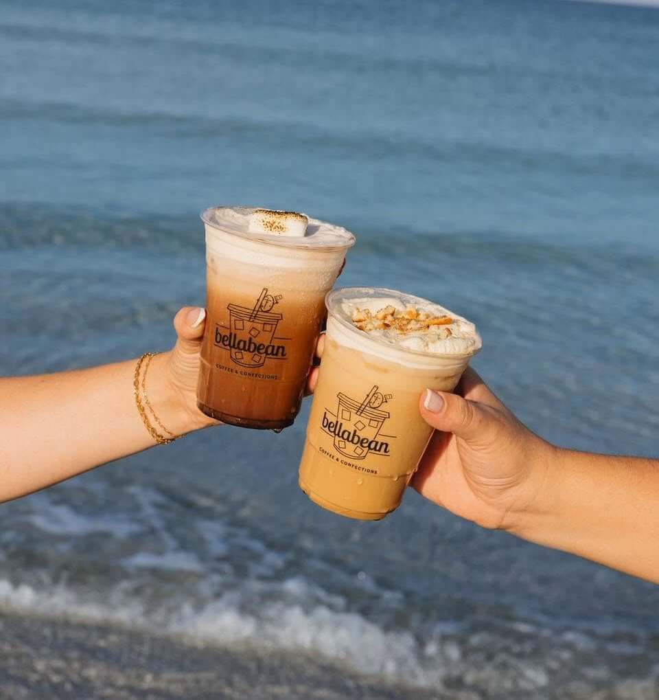 Bradenton Area Coffee Spots two hands holding iced coffee drinks in front of the ocean