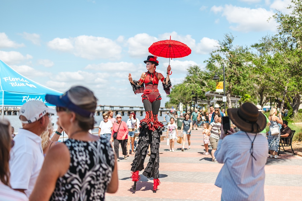 Performer in red and black costume holding red umbrella walking on stilts.
