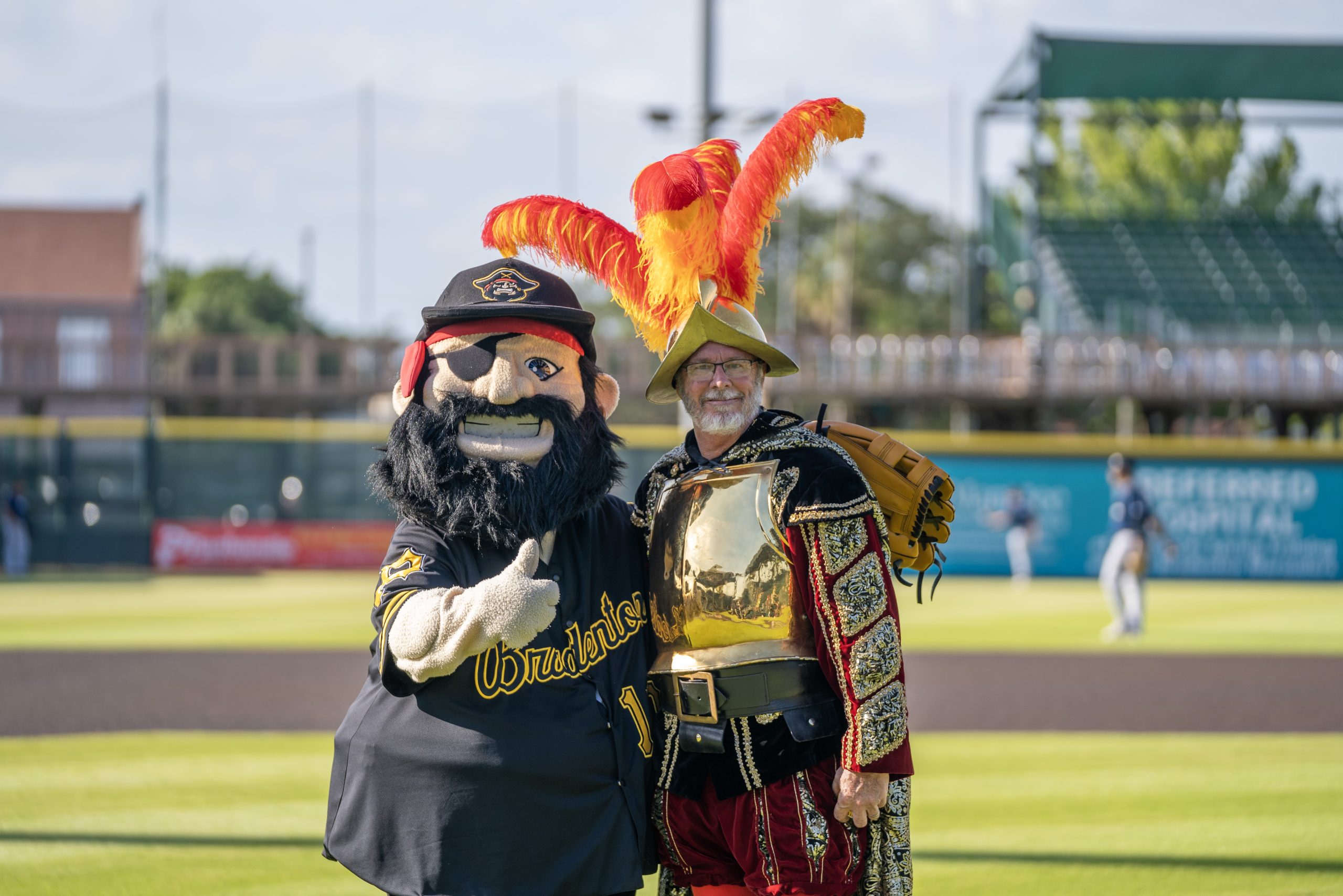 Marty, with thumb up, standing on pitchers mound with a crewe member dressed as a conquistador.