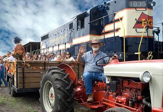 man driver a tractor with a hay ride behind him and a train in the background